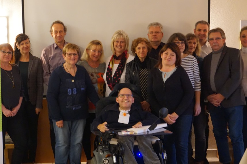 Die Gruppe der kommunalen Behindertenbeauftragten steht zusammen mit dem Landesbehindertenbeauftragten vor einer Leinwand. 