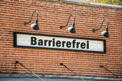 Auf einer Backsteinmauer ist ein beleuchtetes Bild befestigt mit der Aufschrift "Barrierefrei"