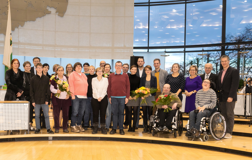 Die Preisträger des 3. Sächsischen Inklusionspreises stehen zusammen mit den Behindertenbeauftragten im Plenarsaal des Sächsischen Landtages als Gruppe zusammen, halten ihre Auszeichnungen und Blumenstäuße in den Händen und lächeln in die Kamera.