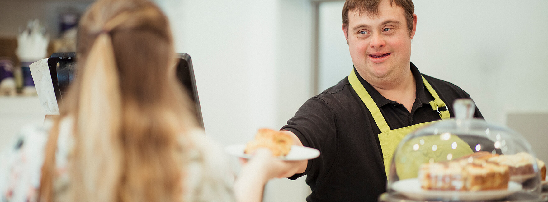 Ein Mann mit Down-Syndrom serviert einer Frau in einem Café, in dem er arbeitet, einen Scone.